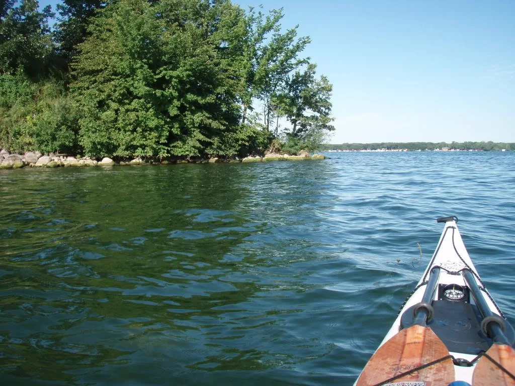 A kayak bow points across rippling blue water beside a tree-covered shoreline.